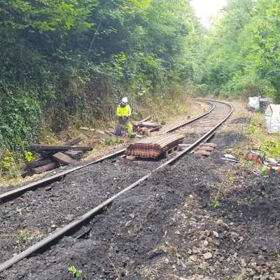 Railroad tracks under maintenance in a forested area with a worker in a yellow vest.