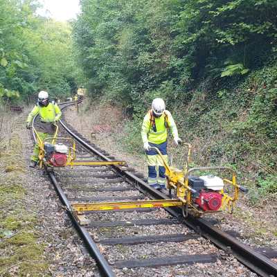 Two workers in protective gear maintaining a railway track in a wooded area.