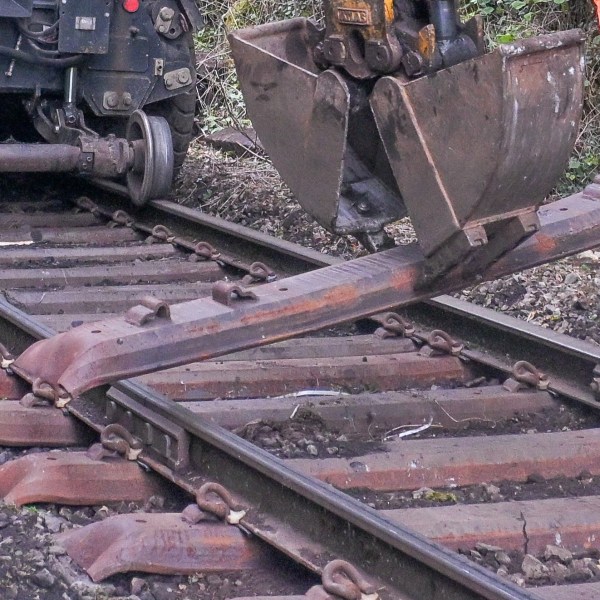 Excavator lifting a railway sleeper near train tracks with vegetation nearby.