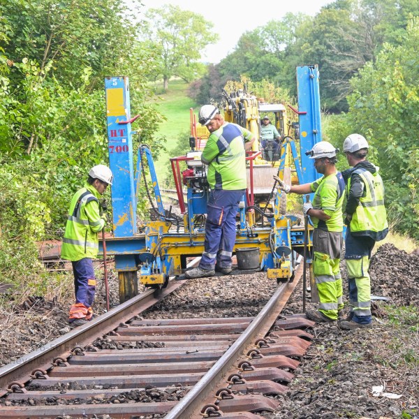 Workers in safety gear operating machinery on railway tracks surrounded by greenery.