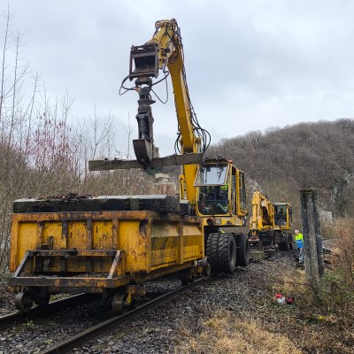 Yellow excavator on railway tracks lifting materials, surrounded by bare trees.