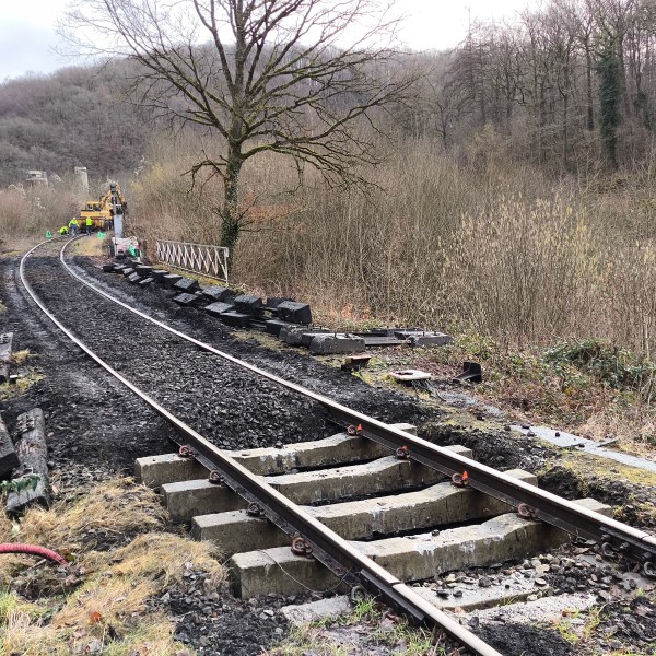 Railway track under construction in a wooded area with machinery and workers in the background.