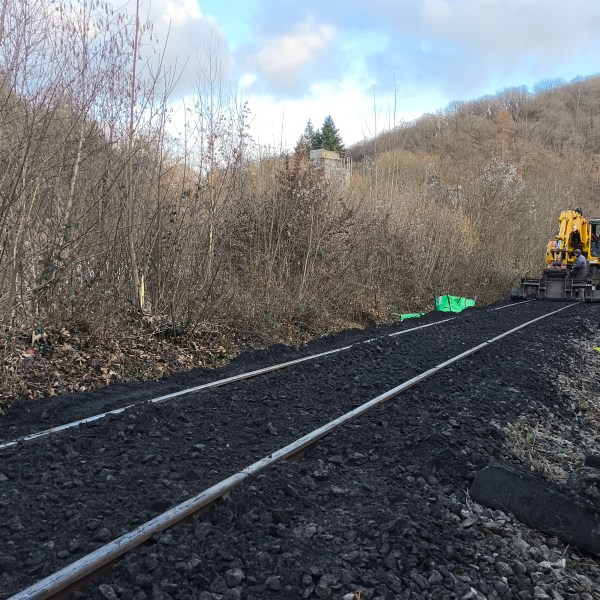 Excavator repairs railway tracks surrounded by barren trees in a hilly landscape.