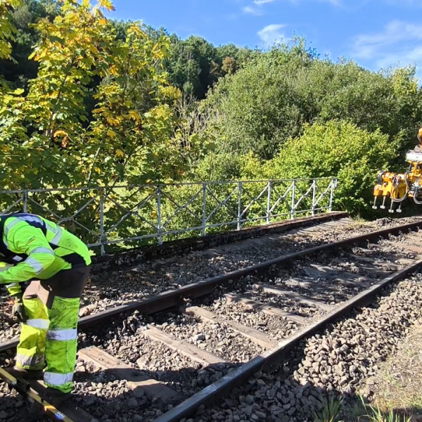 Two workers in safety gear on a railway track with a yellow machine in the background.
