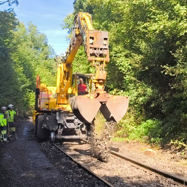 Excavator clearing debris from a railway track, workers observing nearby, surrounded by trees.