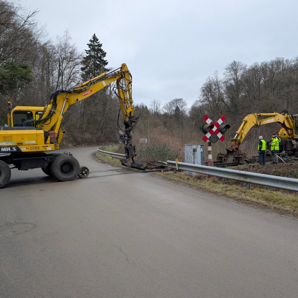 Two construction vehicles near crossing, workers in safety vests, overcast sky with trees in background.