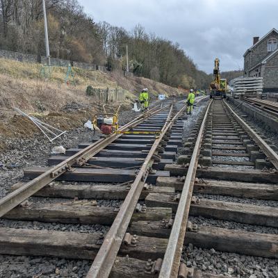 Workers in safety gear repairing train tracks near a building on a cloudy day.