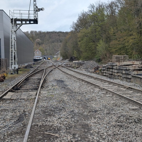 Rail track with switch, beside a building and piles of materials, leading into a wooded area.