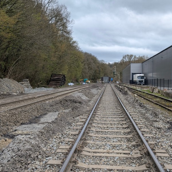Railway tracks with trees on left, industrial building on right, and cloudy sky.