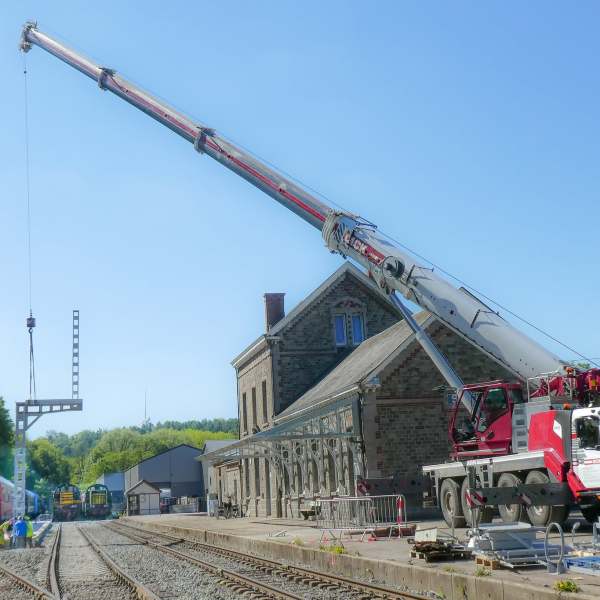 Mobile crane lifting metal structure at train station with tracks and workers.