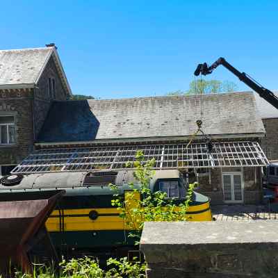 Crane lifts roof structure beside train and stone station building.
