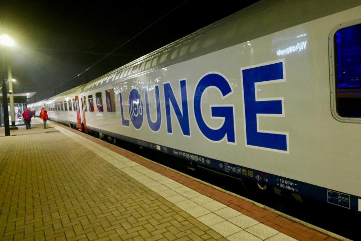 Stationary train with 'LOUNGE' on side at a platform in dim lighting.